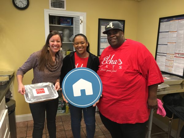 2 women and 1 man smiling; the first woman is holding a covered aluminum tray; the middle woman is holding a blue circle sign with a shelter symbol the man on the left is wearing a red t-shirt;