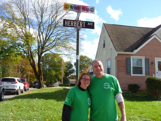 A woman and a man in green t-shirts standing outside on a sunny day under a tie-dye street sign that reads